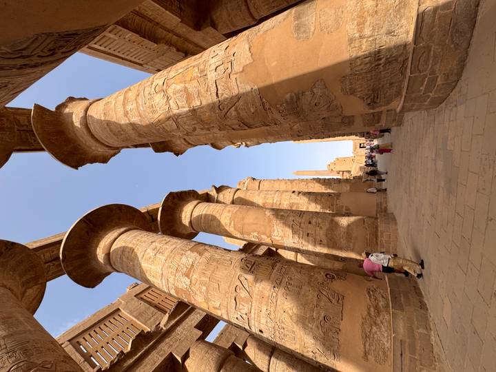 Towering hieroglyph-covered columns line a stone walkway inside a vast Egyptian temple with a few visitors below.