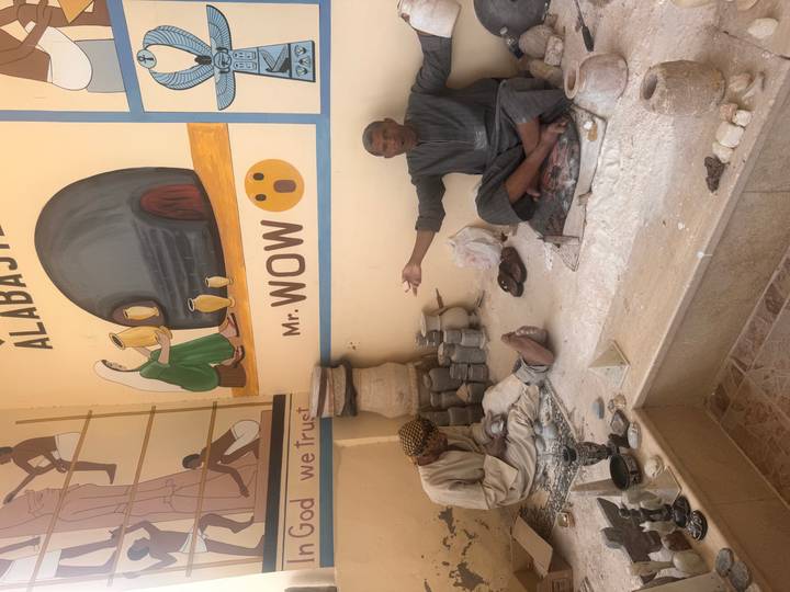 Two craftsmen sit on the floor of an alabaster workshop surrounded by stone powders and carved pieces.