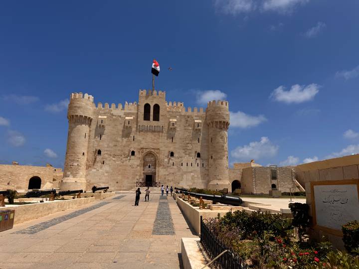 A well-preserved seaside fortress crowned with an Egyptian flag stands under a deep blue sky with scattered visitors below.