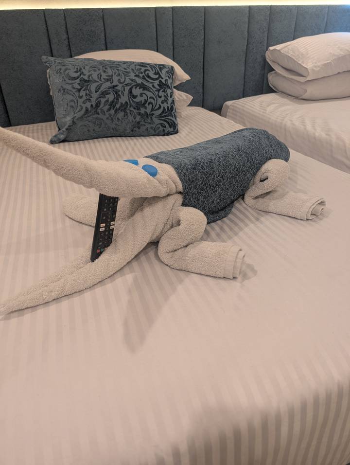 Playful towel animal shaped like a beetle or dinosaur holds a TV remote atop a striped bedspread.