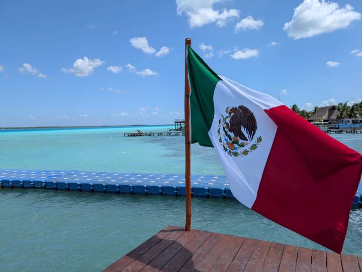Mexican flag waves over the vivid turquoise waters of Bacalar Lagoon under a bright sky.
