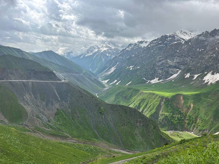 Dramatic alpine valley with winding road, snow-capped peaks and green slopes beneath a partly cloudy sky.