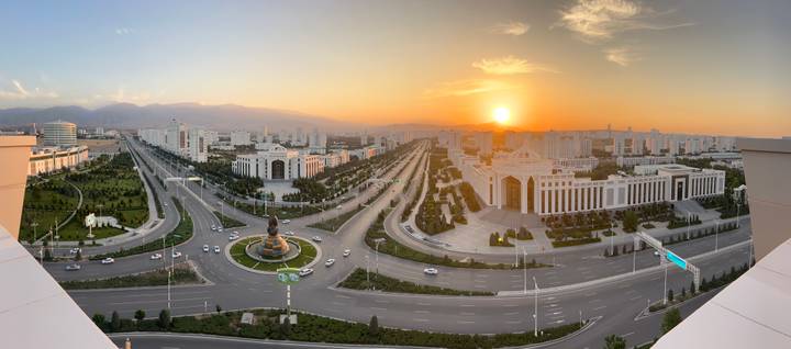 Panoramic sunset view over a grand, marble-clad modern city with broad boulevards and the sun setting behind distant hills.