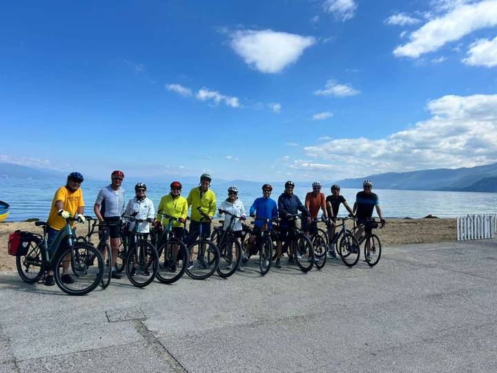 Cycling group lined up with touring bikes beside a calm lakeshore under blue skies.