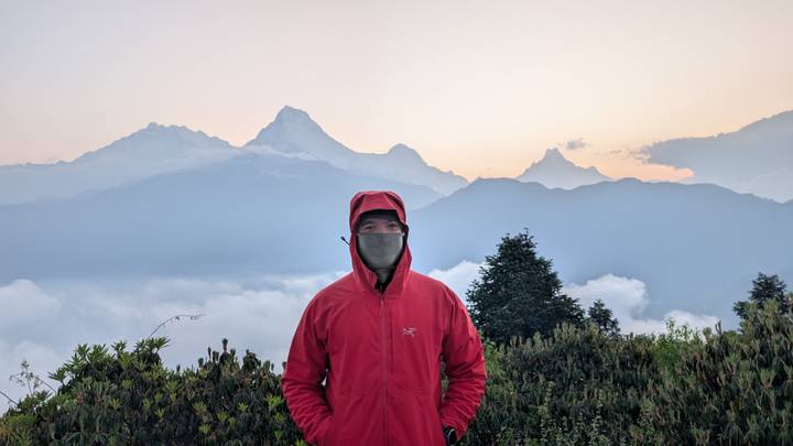 Trekker in red jacket stands before misty Himalayan range at dawn, clouds swirling below.