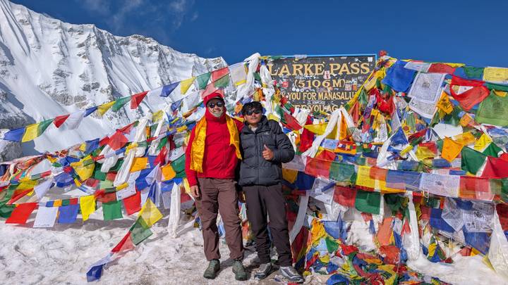 Two trekkers pose at snowy Larke Pass sign surrounded by colorful prayer flags at 5106 m.