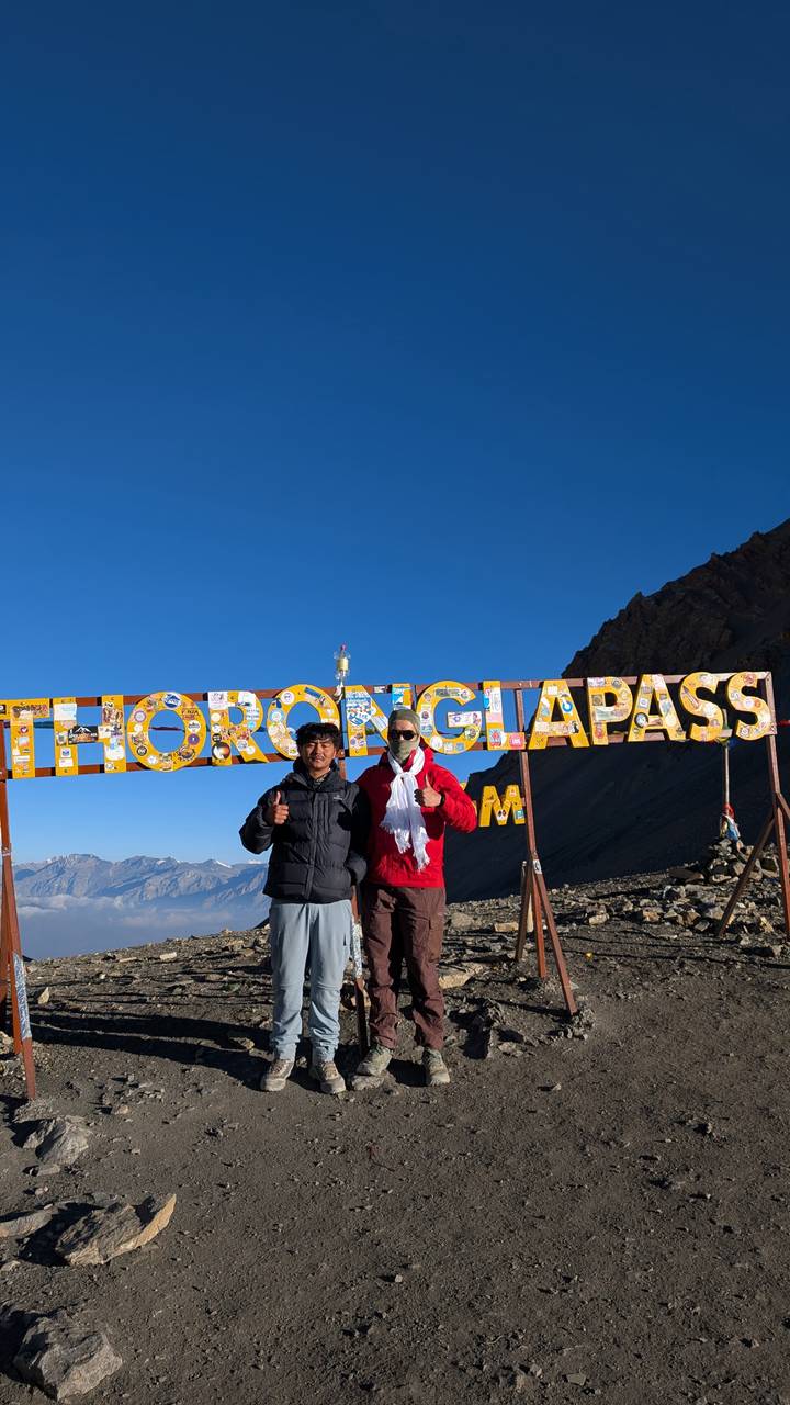 Two trekkers give thumbs-up beneath the colorful THORONG LA PASS summit sign against clear blue sky.