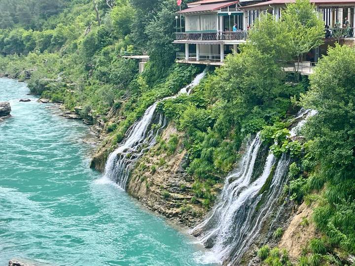 Turquoise river water cascades down multiple small waterfalls beside a cliff topped with a riverside restaurant shaded by trees.