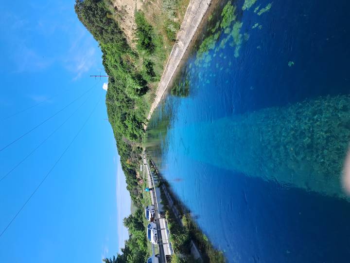 A crystal-clear blue spring forms a calm channel beside a rural roadside under a bright sky.