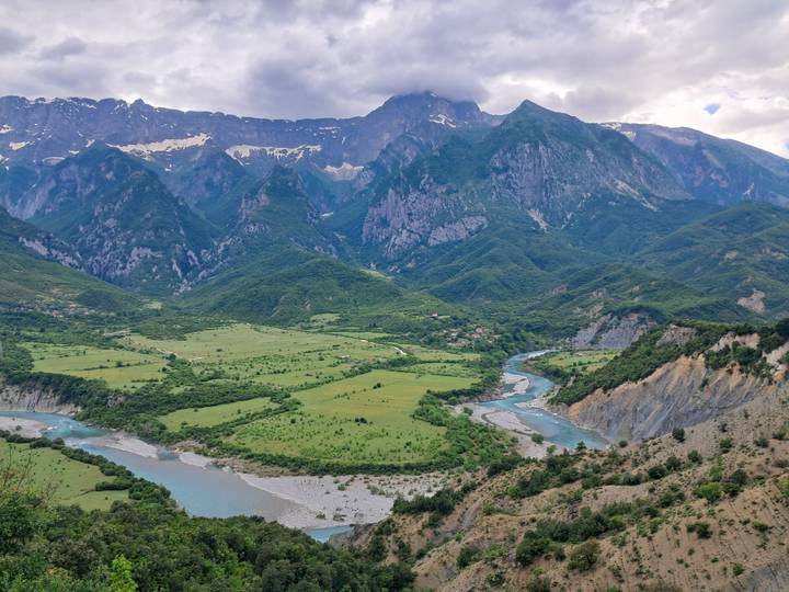 A sweeping alpine valley with a winding blue river sits beneath towering green mountains capped with lingering snow.