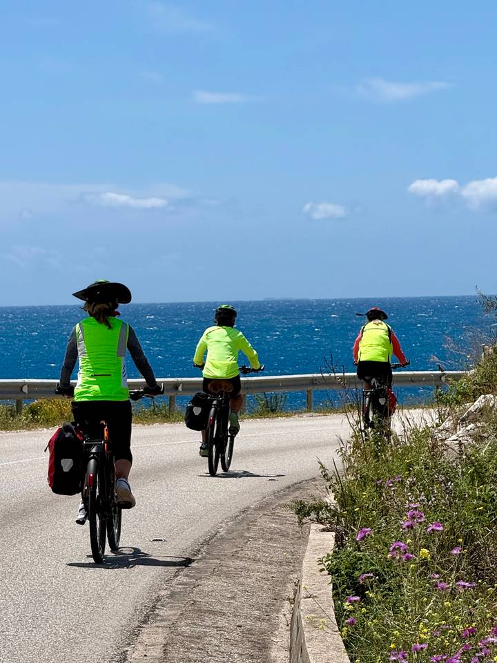 Three cyclists in neon vests ride along a seaside road beside deep blue water.
