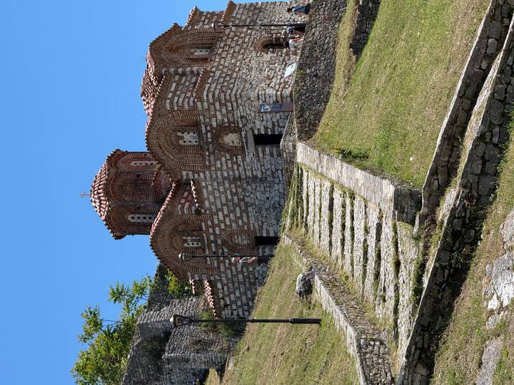 Small medieval stone church with red-tiled dome perched on a grassy slope accessed by stone steps.