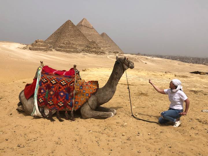 Traveler gestures to a seated camel with pyramids rising behind in sandy landscape