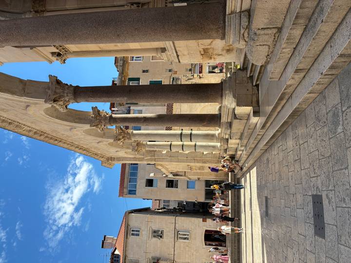 Sunlit Roman columns and arches of Split's Peristyle square with visitors relaxing on the steps.