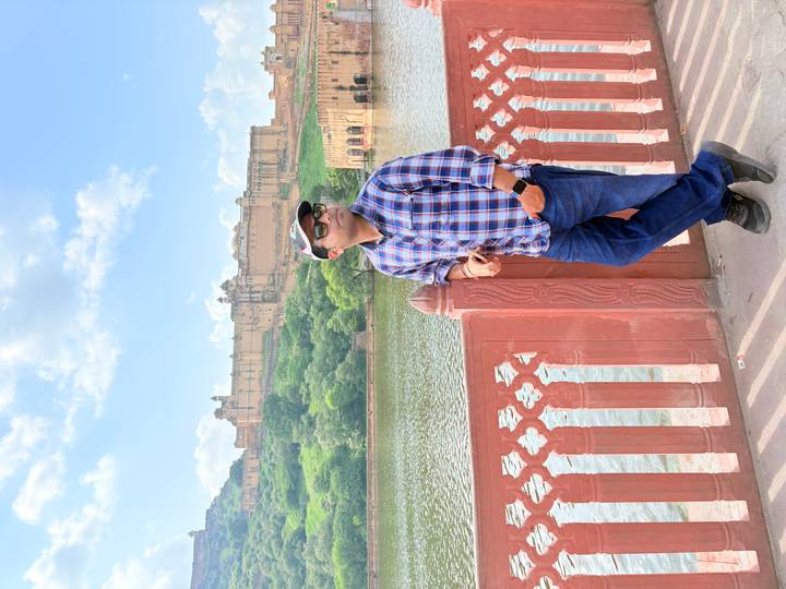 Traveler poses by the lakeside railing with Amber Fort rising on the opposite hill under a blue sky.