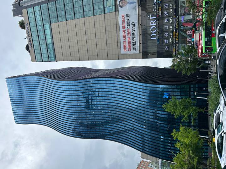 Tall wavy blue-glass skyscraper rising next to a beige office block under grey sky in Seoul