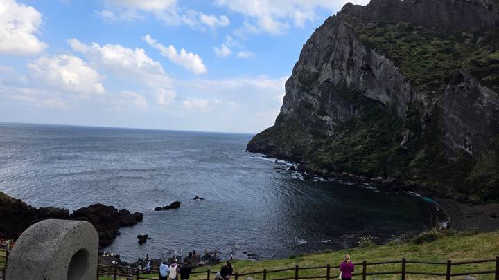 Volcanic cliff and bay on Jeju Island with visitors gathered along the shoreline.