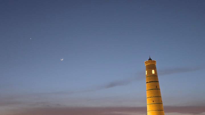Slim illuminated minaret reaches into a deep twilight sky beside a crescent moon.