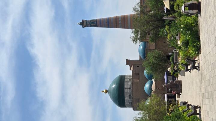 View across gardens toward turquoise-domed mosque and striped minaret inside Khiva’s old town.