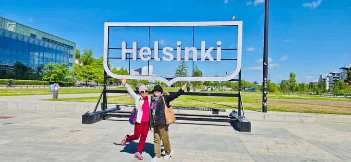 Two smiling women posing under a large outdoor Helsinki sign on sunny day.