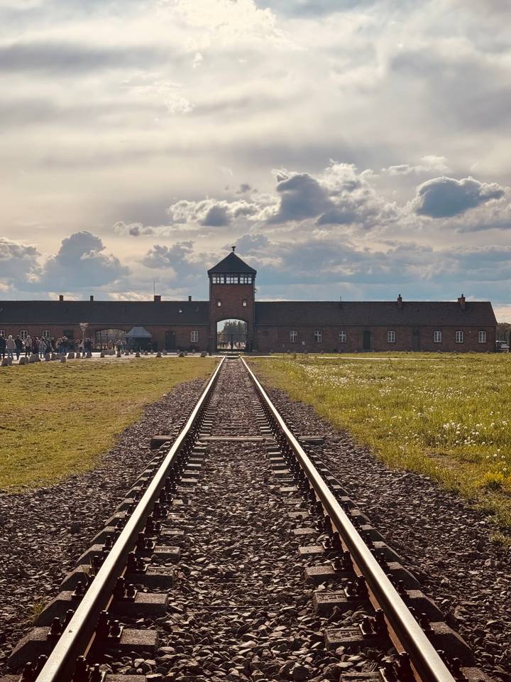 Iconic railway tracks leading to brick entrance gate of Auschwitz-Birkenau under dramatic sky.