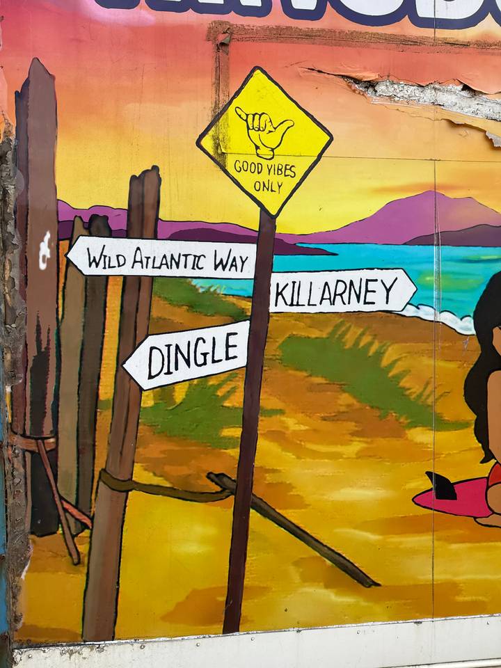 Colorful mural-style signposts pointing toward Wild Atlantic Way, Killarney and Dingle on a painted coastal background.