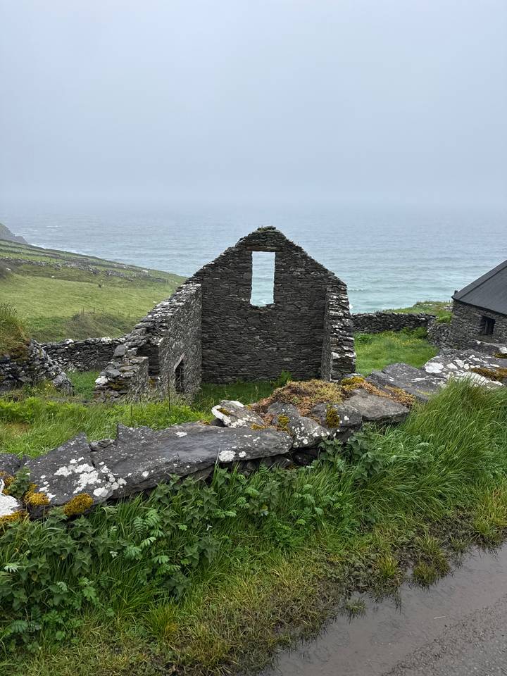 Stone ruin of a roofless cottage overlooking green cliffs and the Atlantic Ocean under misty skies.