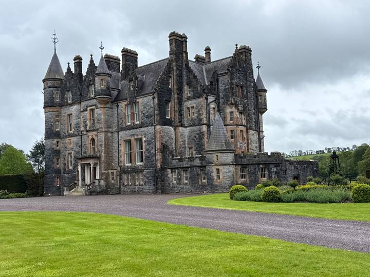 Grand gothic-style stone castle with turrets and manicured lawn under grey clouds.