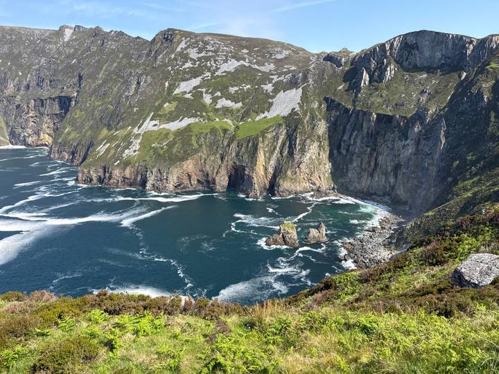 Dramatic high sea cliffs covered with green patches dropping into swirling blue Atlantic waters.