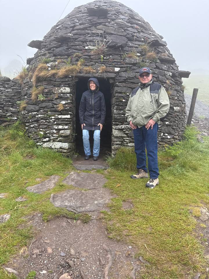 Two travelers at the doorway of an ancient beehive stone hut on a misty hillside.