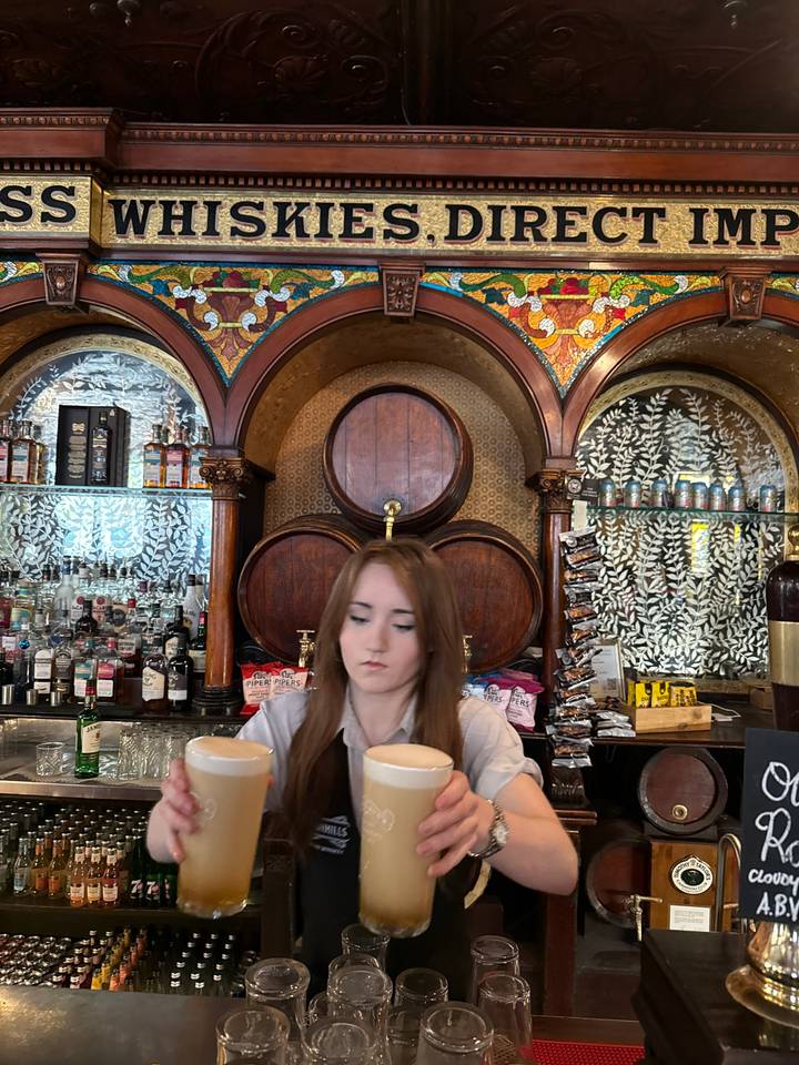 Bartender pouring a drink behind an ornate wooden bar with shelves of spirits.