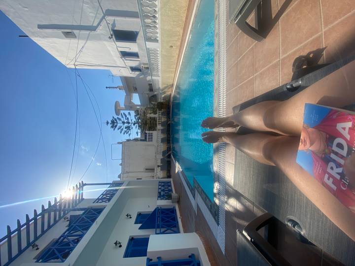 Relaxing poolside view with sun-bathing traveller’s legs, turquoise water and bright Greek apartments.