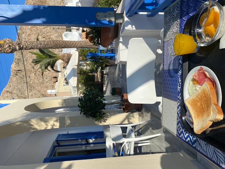 Outdoor breakfast setting on a sunny terrace with white and blue Cycladic architecture, palm tree and rocky hill in the background, toast, ham, egg and orange juice on a tray.