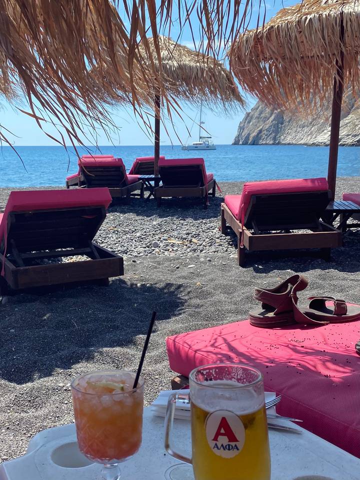 Row of pink-cushioned sun-loungers on a pebbly black-sand beach facing calm blue sea with a boat offshore and sandals on the sand.