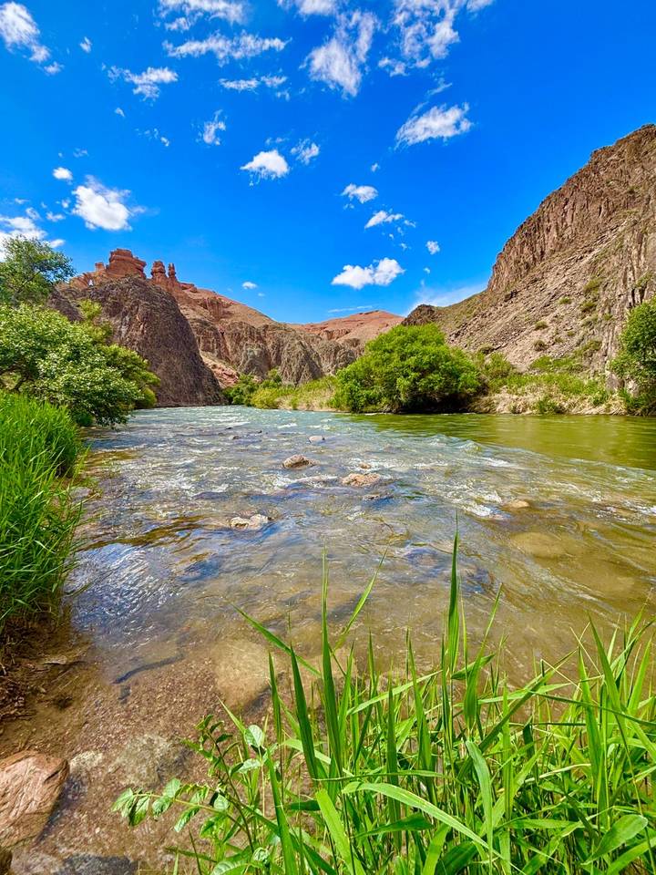 Clear mountain river rushing between green banks and red sandstone canyon walls beneath bright blue sky.