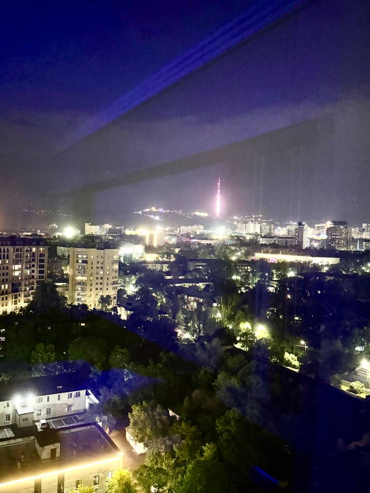 Blurry night view over city lights with glowing pink TV tower in the distance framed by dark buildings.