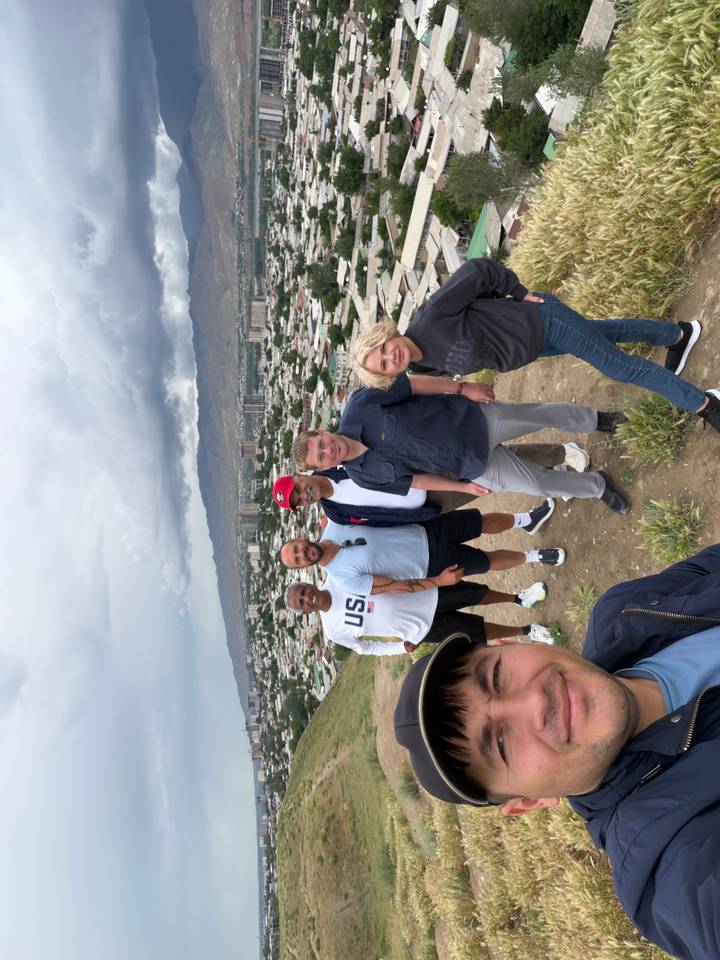 Six travelers pose on a hillside viewpoint overlooking a large city and distant mountains under a cloudy sky.