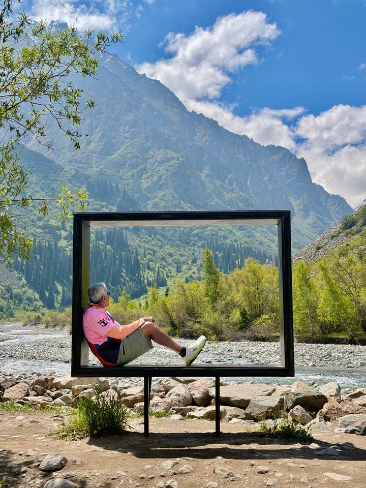 A man relaxes inside a large rectangular frame beside a clear river with lush green mountains towering behind.