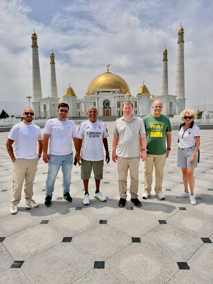 Travelers stand in front of a grand white-and-gold mosque with domes and minarets on a bright day.