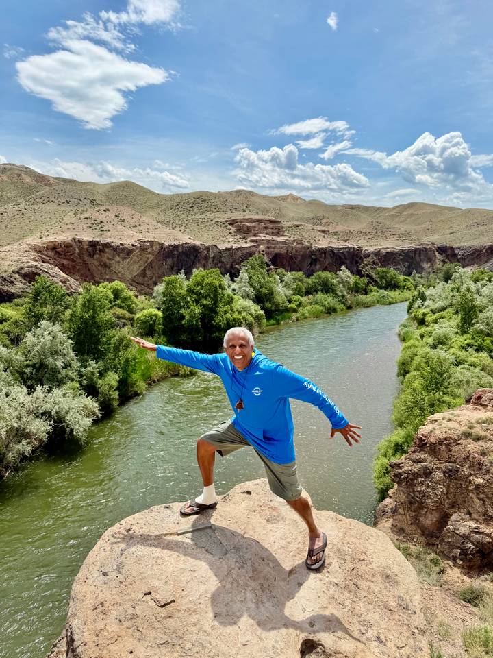 A man in a bright blue hoodie strikes a playful pose on a cliff edge above a green river and desert hills.