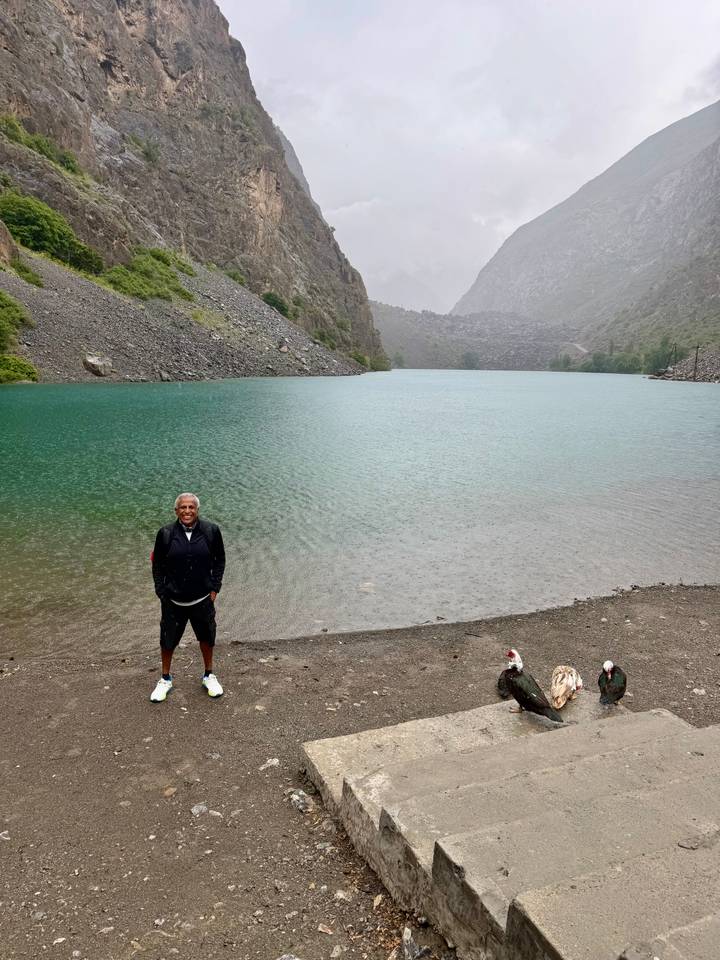 A lone traveler stands beside a turquoise alpine lake surrounded by rocky slopes under misty skies.