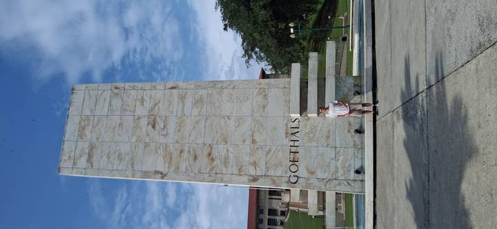 Traveler standing in front of the tall marble Goethals Monument against a partly cloudy sky.