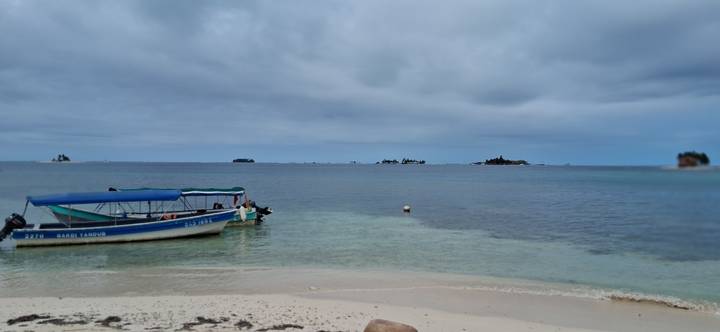 Small motorboat anchored in shallow clear water off a sandy shore with low islands on the horizon under grey clouds.