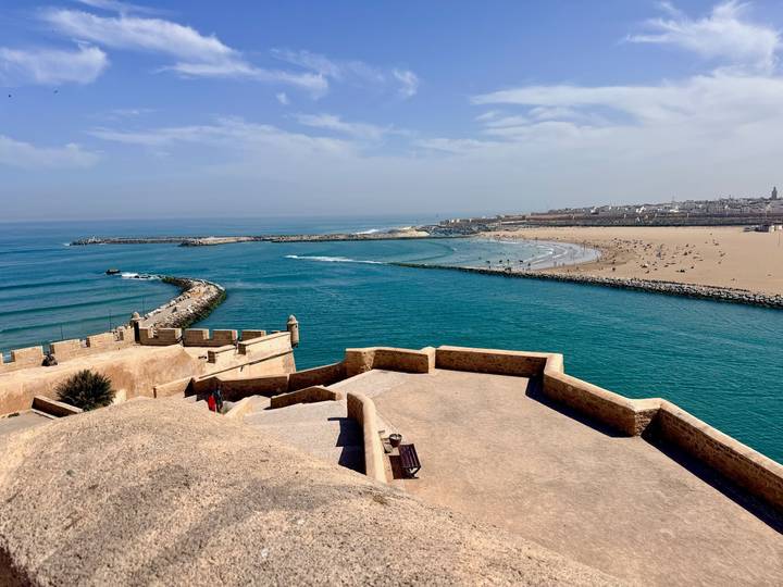 Azure Atlantic waters curve into a sandy bay beside fortress ramparts and a distant town on Morocco’s coast