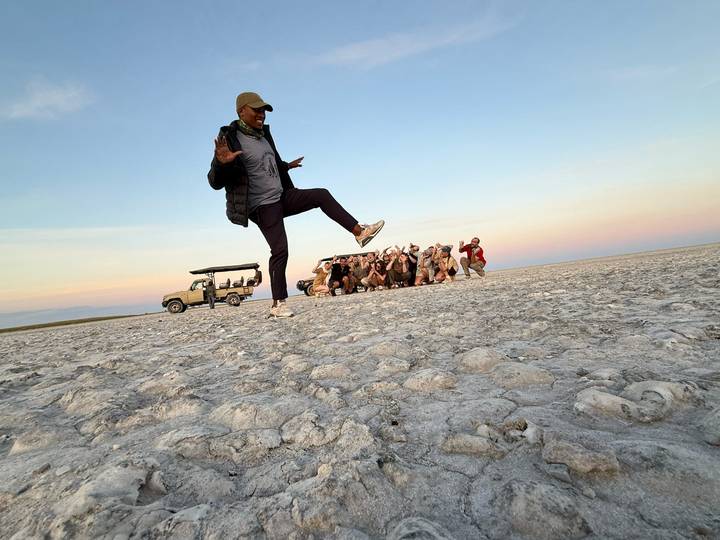 Guide playfully poses while group crouches behind jeep on expansive salt flat at dusk