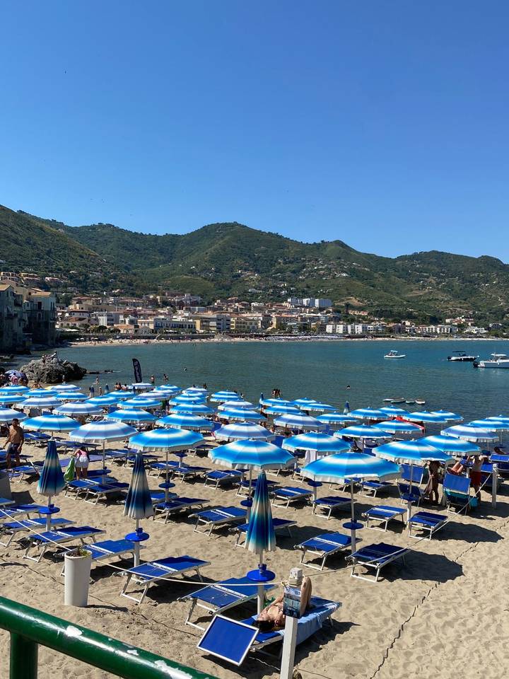 Mediterranean beach scene with rows of blue-striped umbrellas, boats offshore, and green hills behind a coastal town.