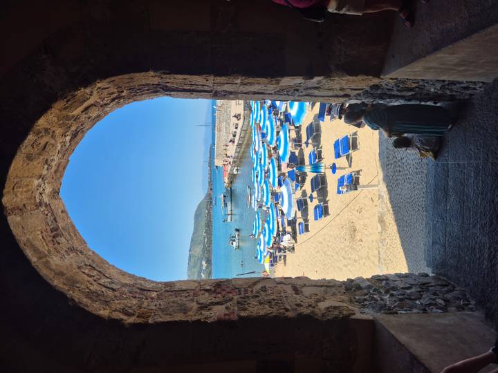 Beach view framed by a stone archway, revealing blue umbrellas and sun loungers on golden sand.