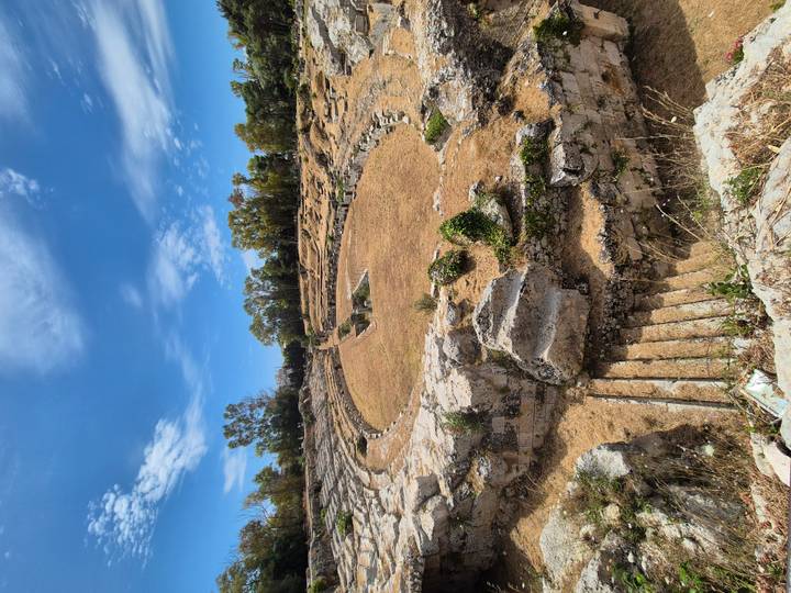 Ruins of a stone amphitheatre ring with stepped seating and central arena surrounded by dry grass.