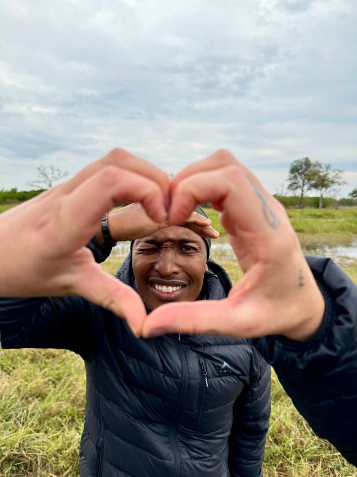 Playful close-up portrait of a man framed by hands forming a heart shape with grassy wetlands behind.