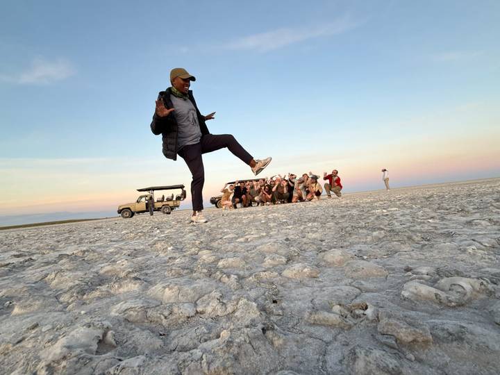 Safari group crouches in the distance while a guide playfully kicks in the foreground on a vast salt pan at sunset.